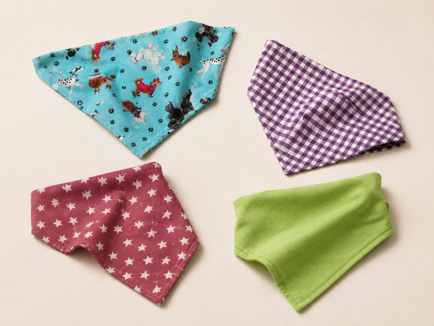 Four Cooper Project dog bandanas laid flat on sand: turquoise with a dog print, purple gingham, dusky pink with white stars, and plain lime green
