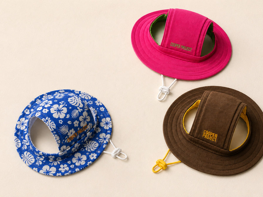 Three Cooper Project dog bucket hats laid flat on sand: pink with a green lining, blue hibiscus print, and brown with a yellow lining, each with a drawcord and COOPER PROJECT embroidered on the crown